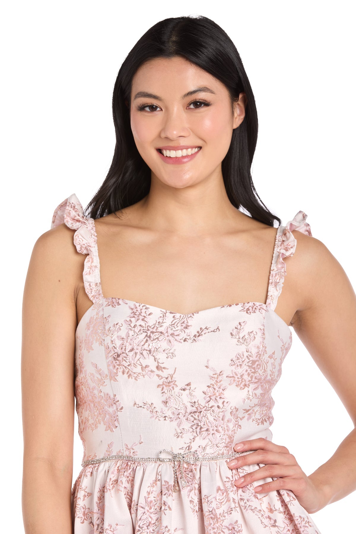 Woman smiling and posing in the Short Pink Print Party Dress: Morgan 14040, a sleeveless floral dress with ruffled straps and a decorative belt, against a white background.